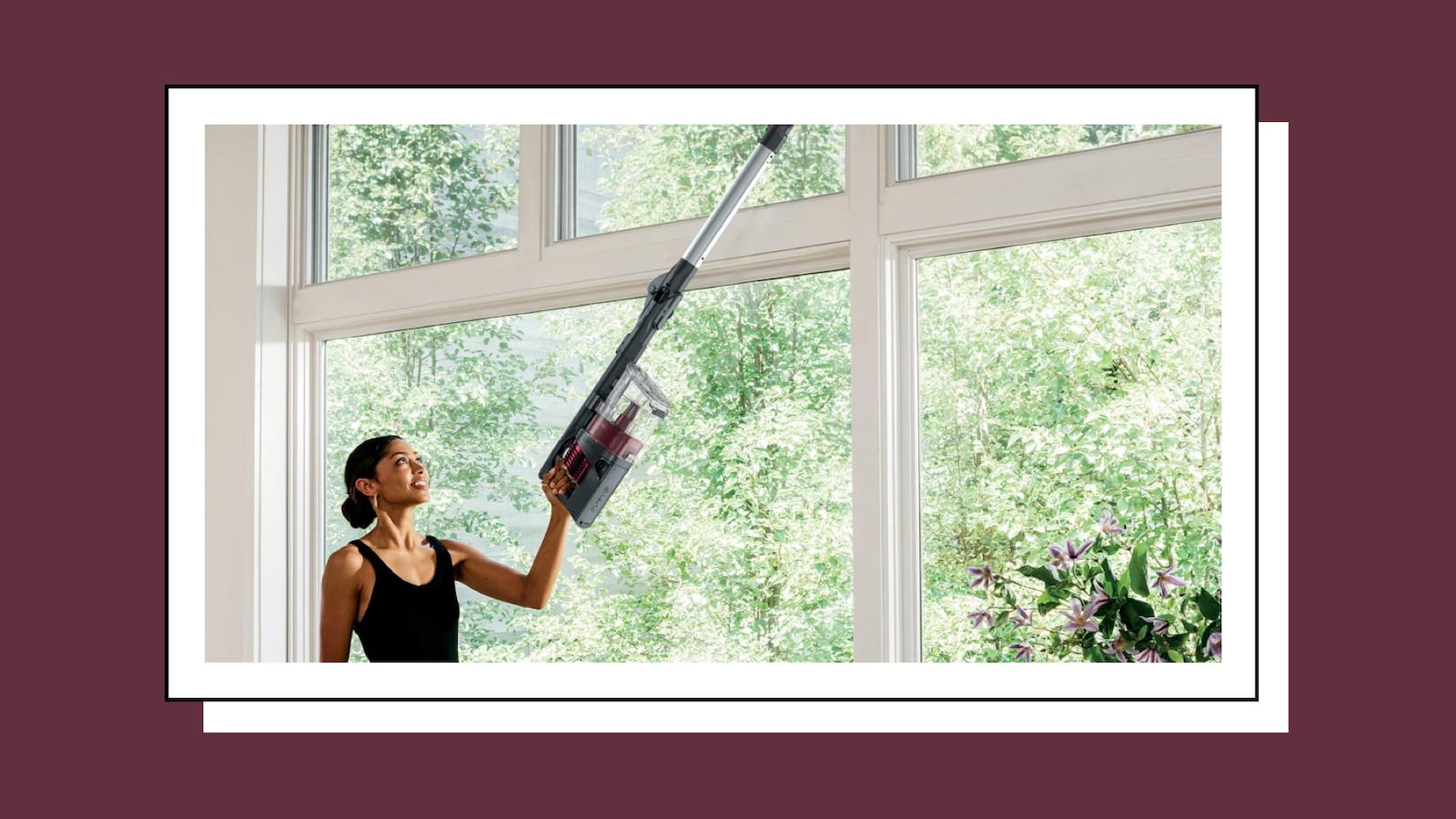 A woman using a cordless vacuum to clean her window sills. She's reaching a sill above her head.