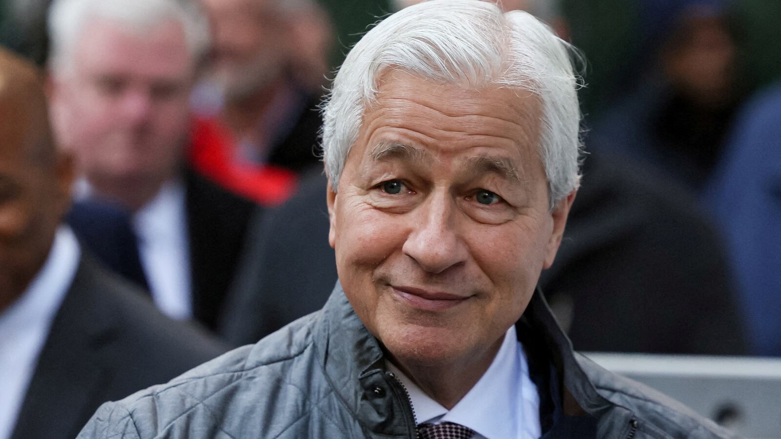 Jamie Dimon waves his hand and smirks outdoors.