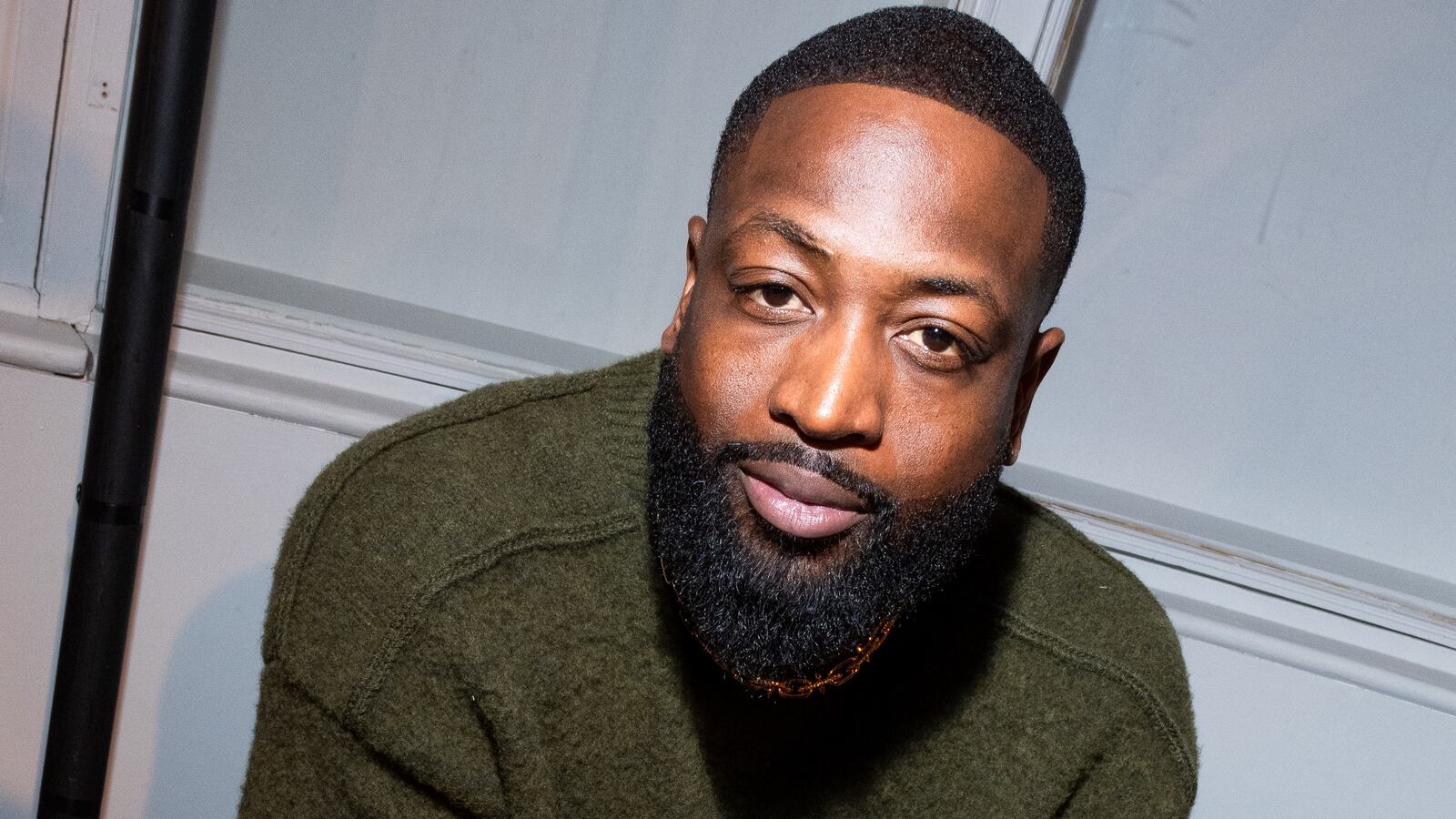 Dwyane Wade sits on a chair alone with his hands clasped together in a green sweater.