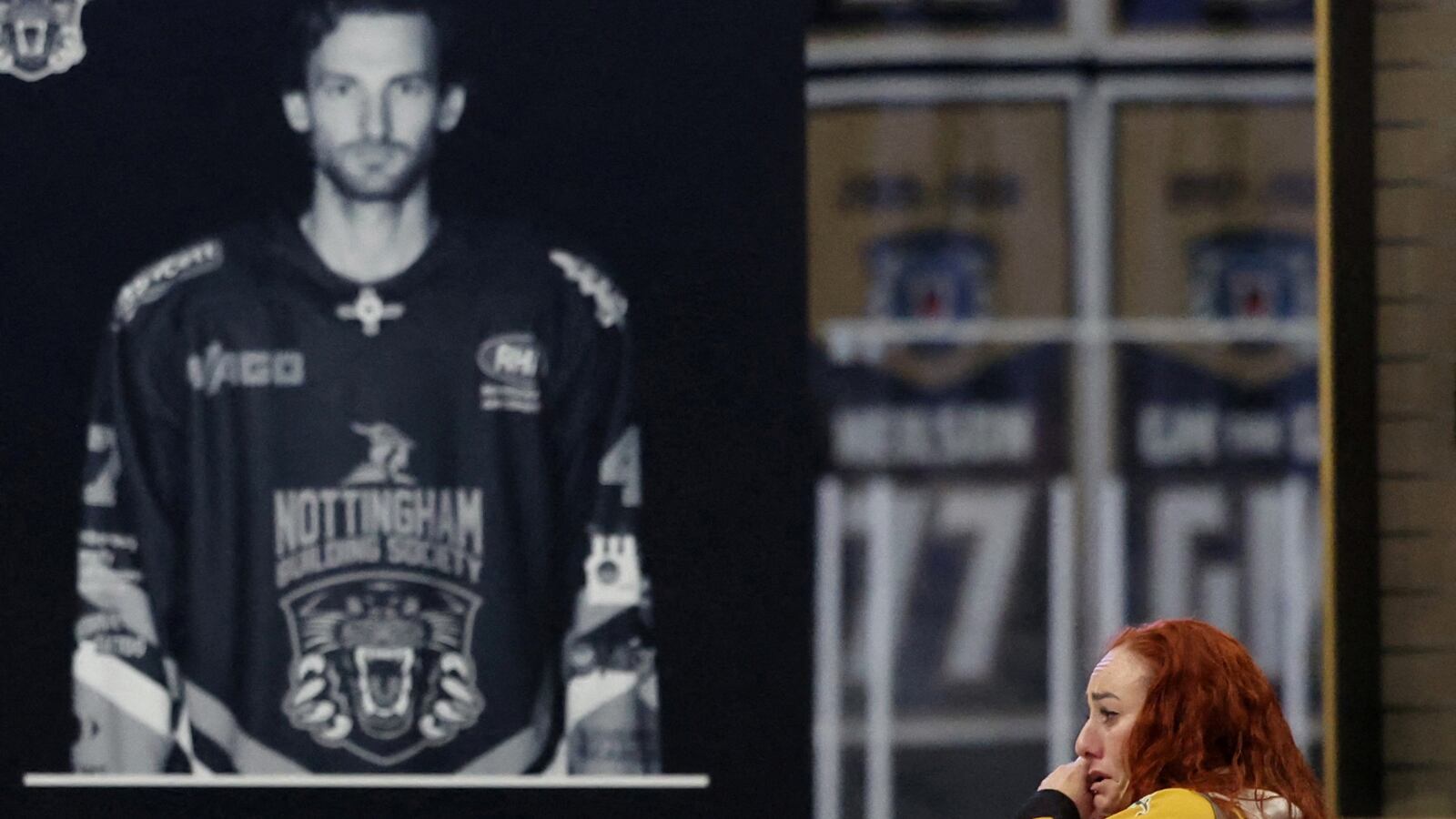 A banner of Matt Johnson is hung as a woman wearing a hockey jersey cries in the foreground.