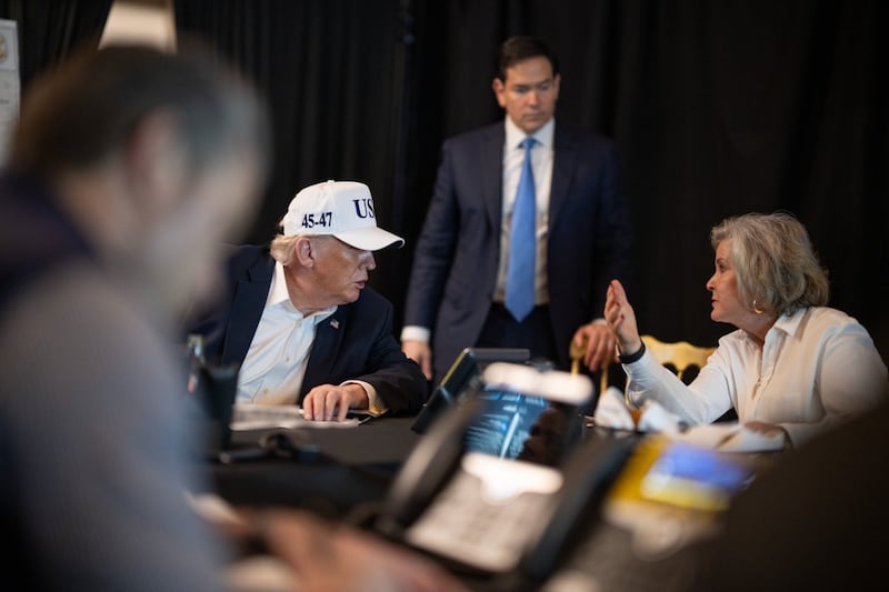Trump in a white USA trucker hat looking at Susie Wiles with Marco Rubio behind him.