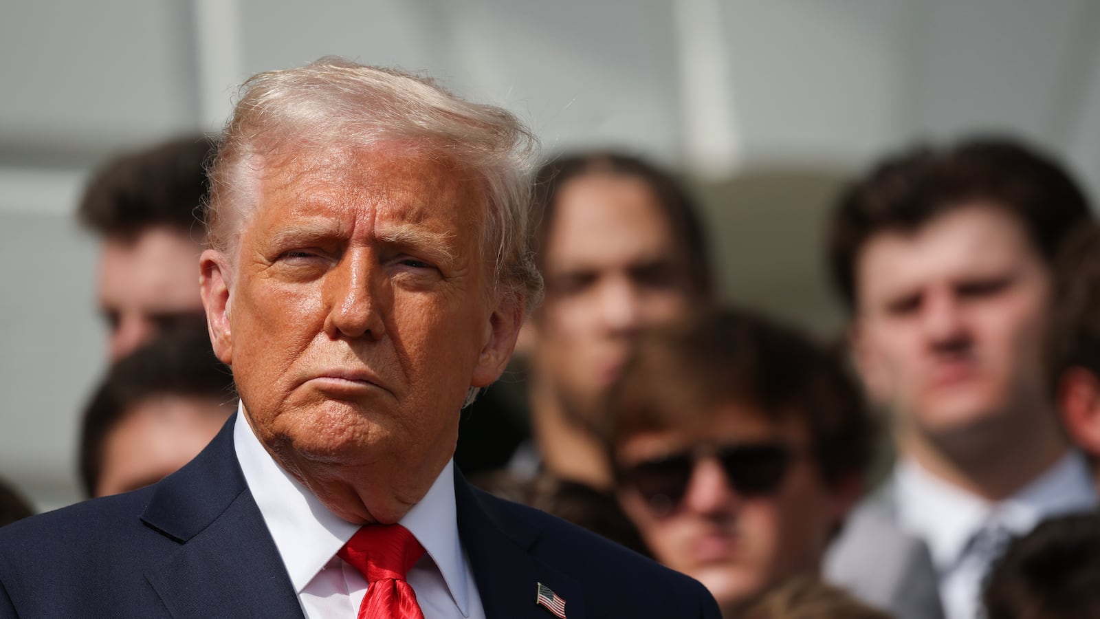 A close-up of President Donald Trump in a suit and tie with football players behind him.