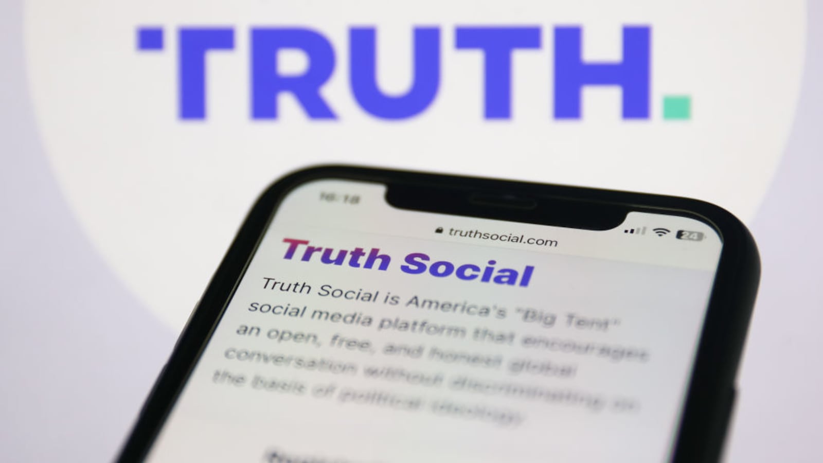 Truth Social website is seen displayed on a phone screen and the Truth Social logo is displayed on a screen behind it.
