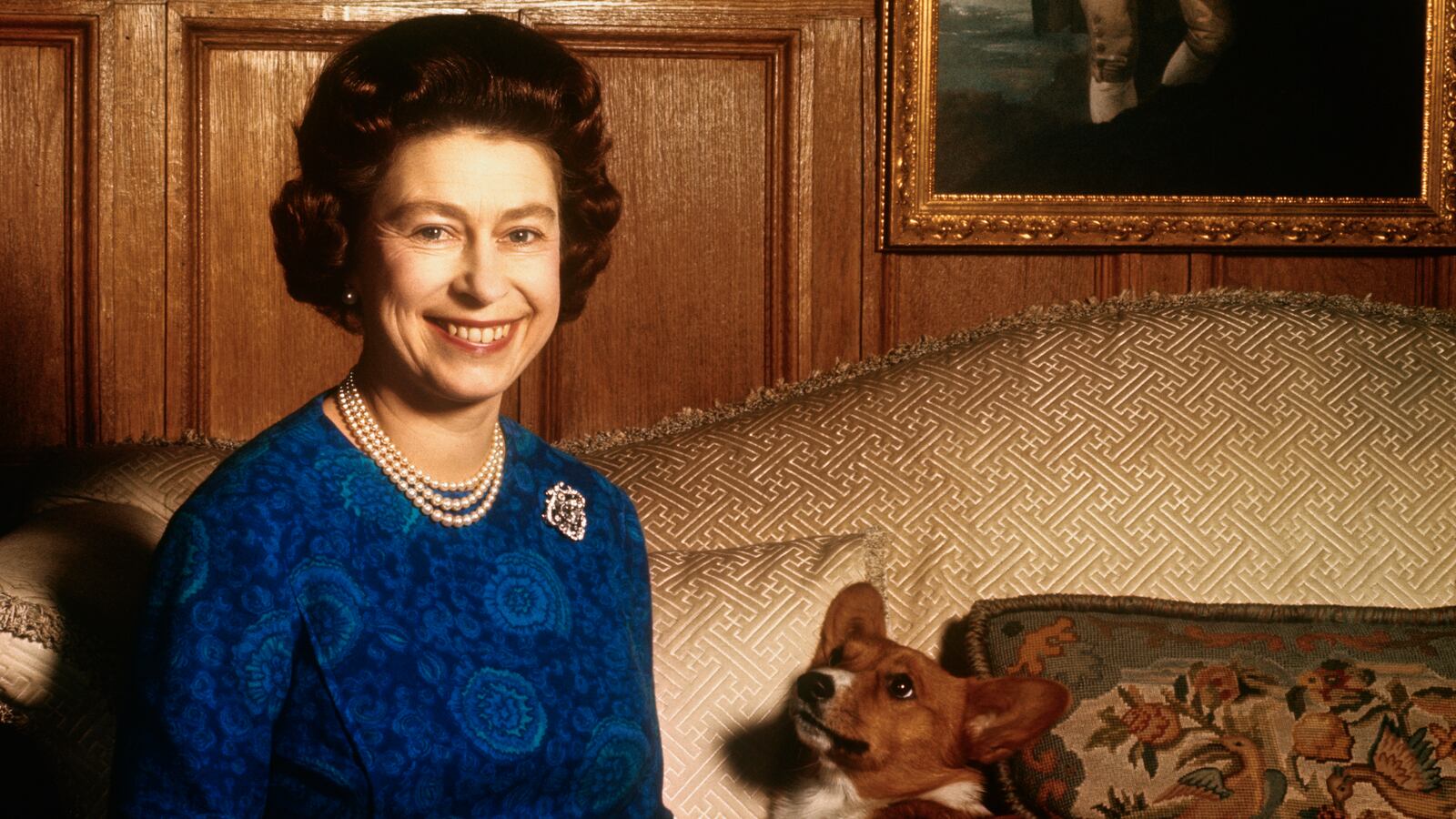 Queen Elizabeth II smiles radiantly during a picture-taking session in the salon at Sandringham House. Her pet dog looks up at her.