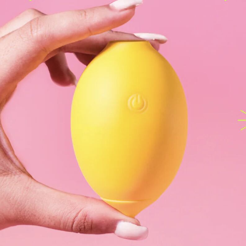 Hand holding a yellow lemon-shaped clitoral massager from Nancy against a pink background.
