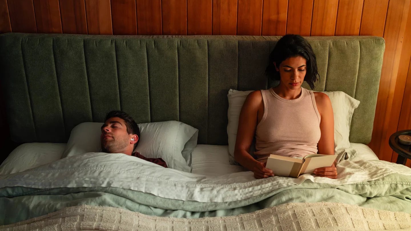 A man wearing Ozlo Sleepbuds asleep in a bed next to a woman reading a book