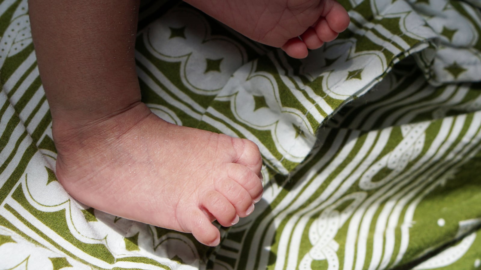 A heartwarming close-up shot focusing on the delicate, wrinkled feet of a sleeping infant, resting comfortably on a richly patterned fabric of olive green and white stripes and clover-like designs und