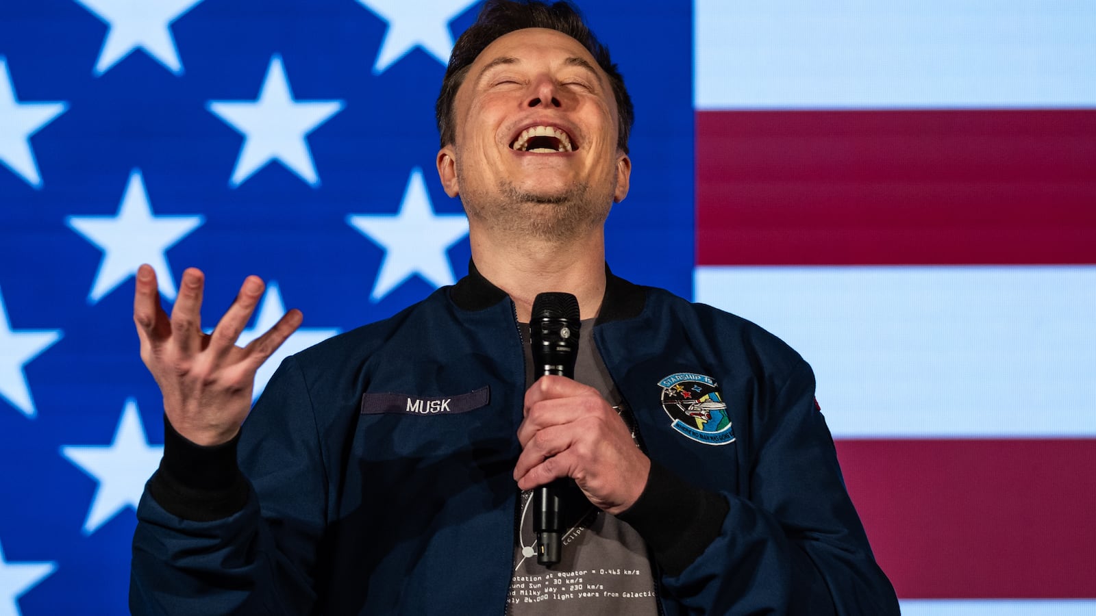 Elon Musk looking up and grinning in front of an American flag.