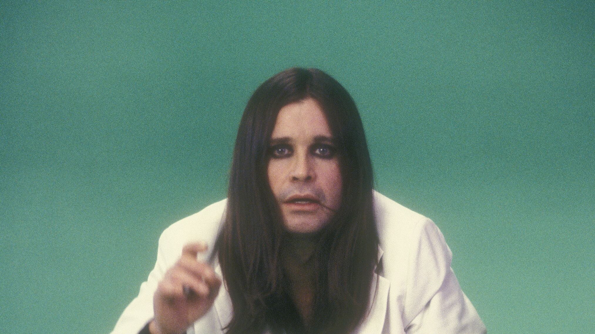 a vintage photo of rock star Ozzy Osbourne in a white suit with long straight black hair and black eyeshadow