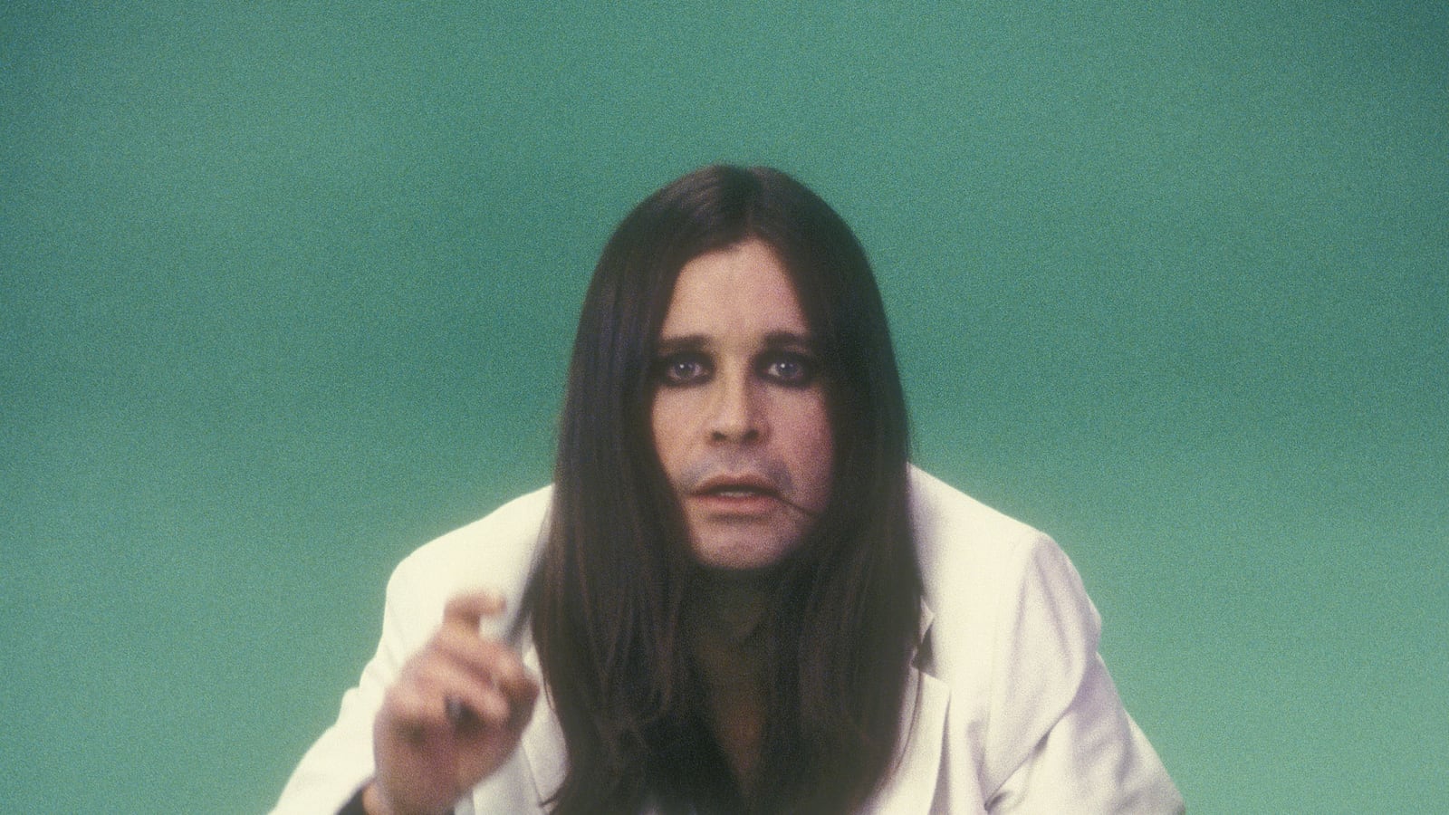 a vintage photo of rock star Ozzy Osbourne in a white suit with long straight black hair and black eyeshadow