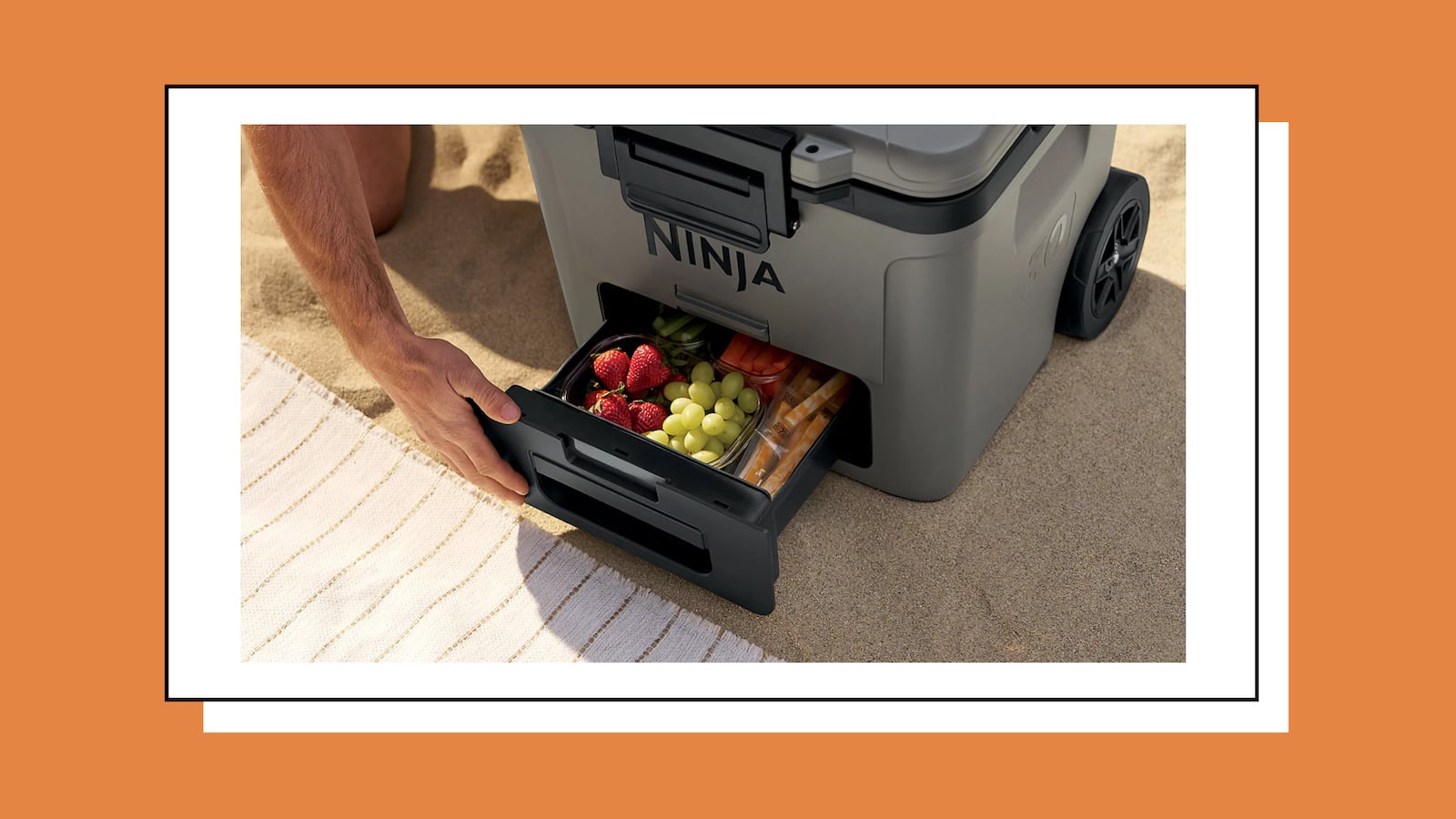 Person pulling open the bottom Dry Zone drawer of a gray Ninja FrostVault wheeled cooler on a sandy beach, revealing compartments filled with strawberries, grapes, and snacks