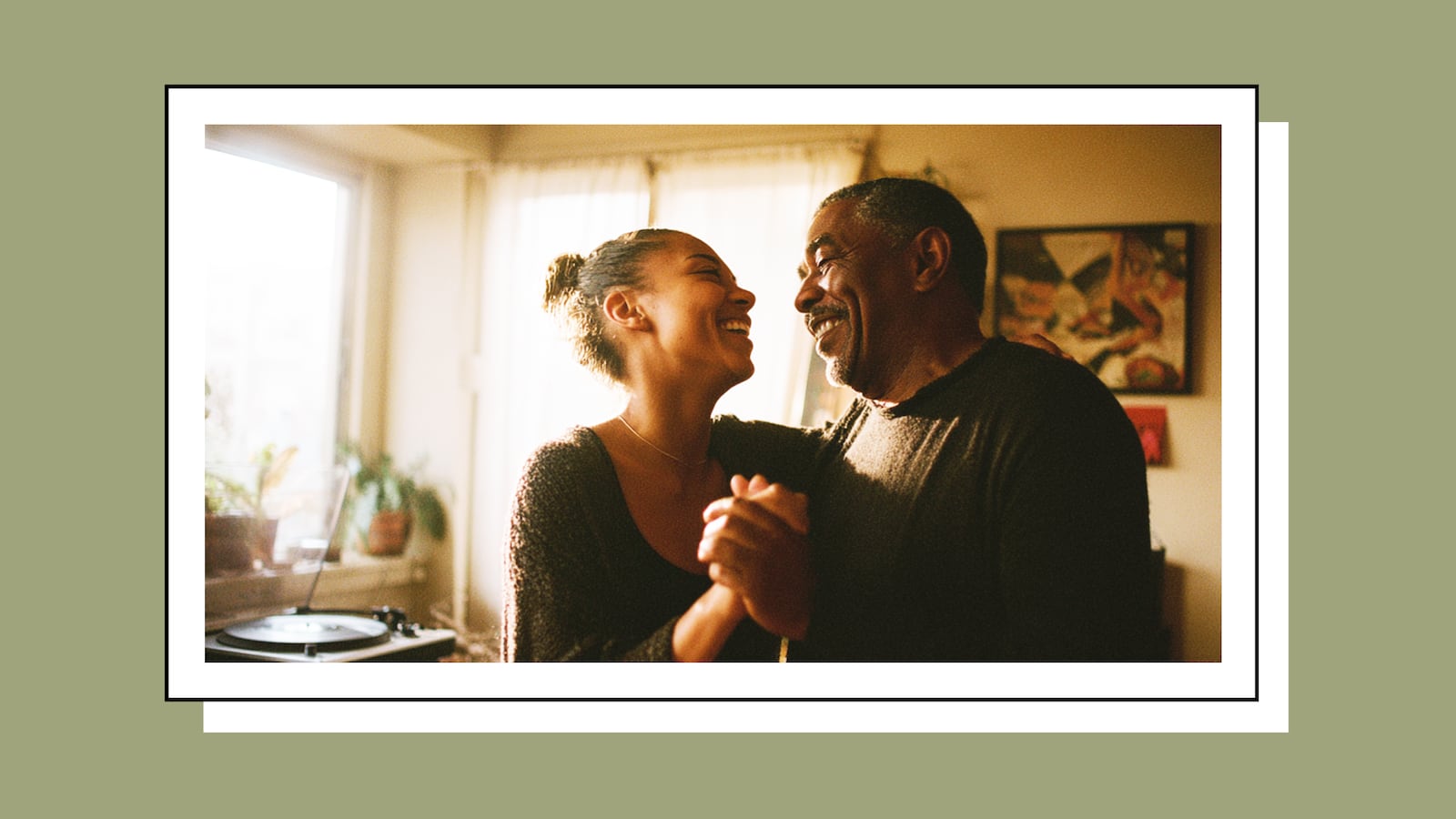 A couple dances together in a warmly lit living room, laughing and holding hands, with a vinyl record player visible in the background.
