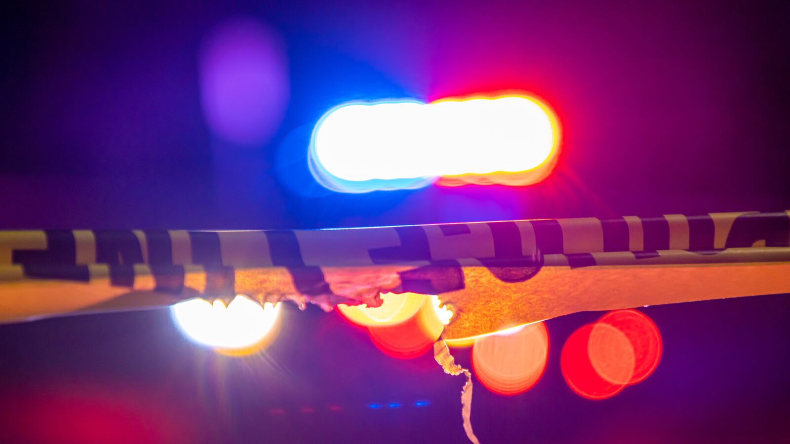 A defocused police car sits behind crime scene tape with flashing lights at night.