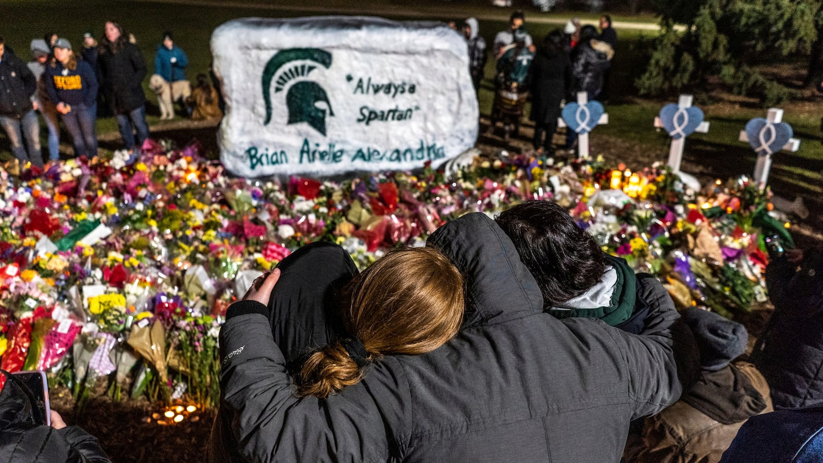 People comfort each other amongst bouquets of flowers during a vigil at The Rock on the Michigan State University campus.