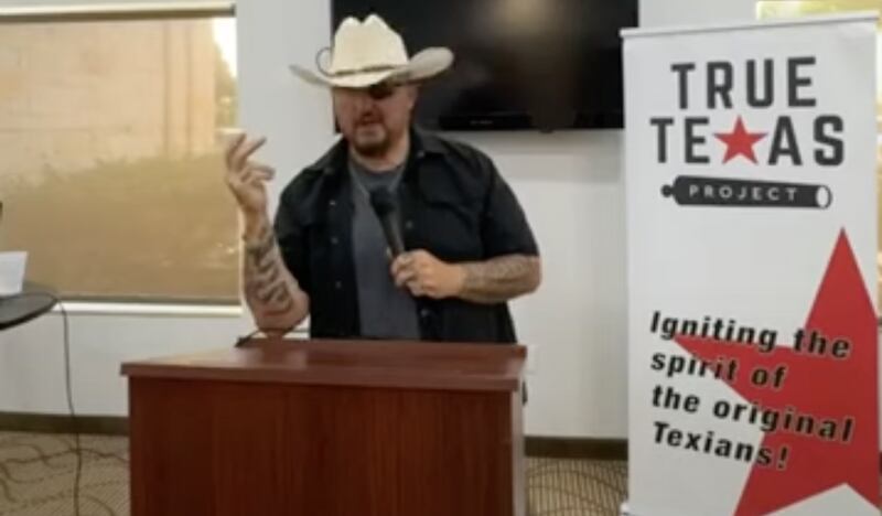 A man in a cowboy hat at a lectern. Beside him is a poster saying "True Texas Project. Igniting the spirit of the original Texians."