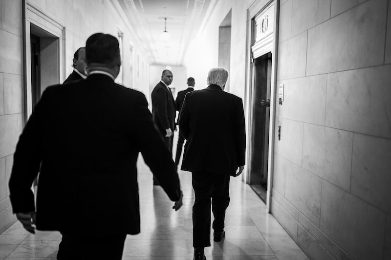 Trump in black and white with this back to the camera walking towards a door in a corridor, surrounded by men in suits.
