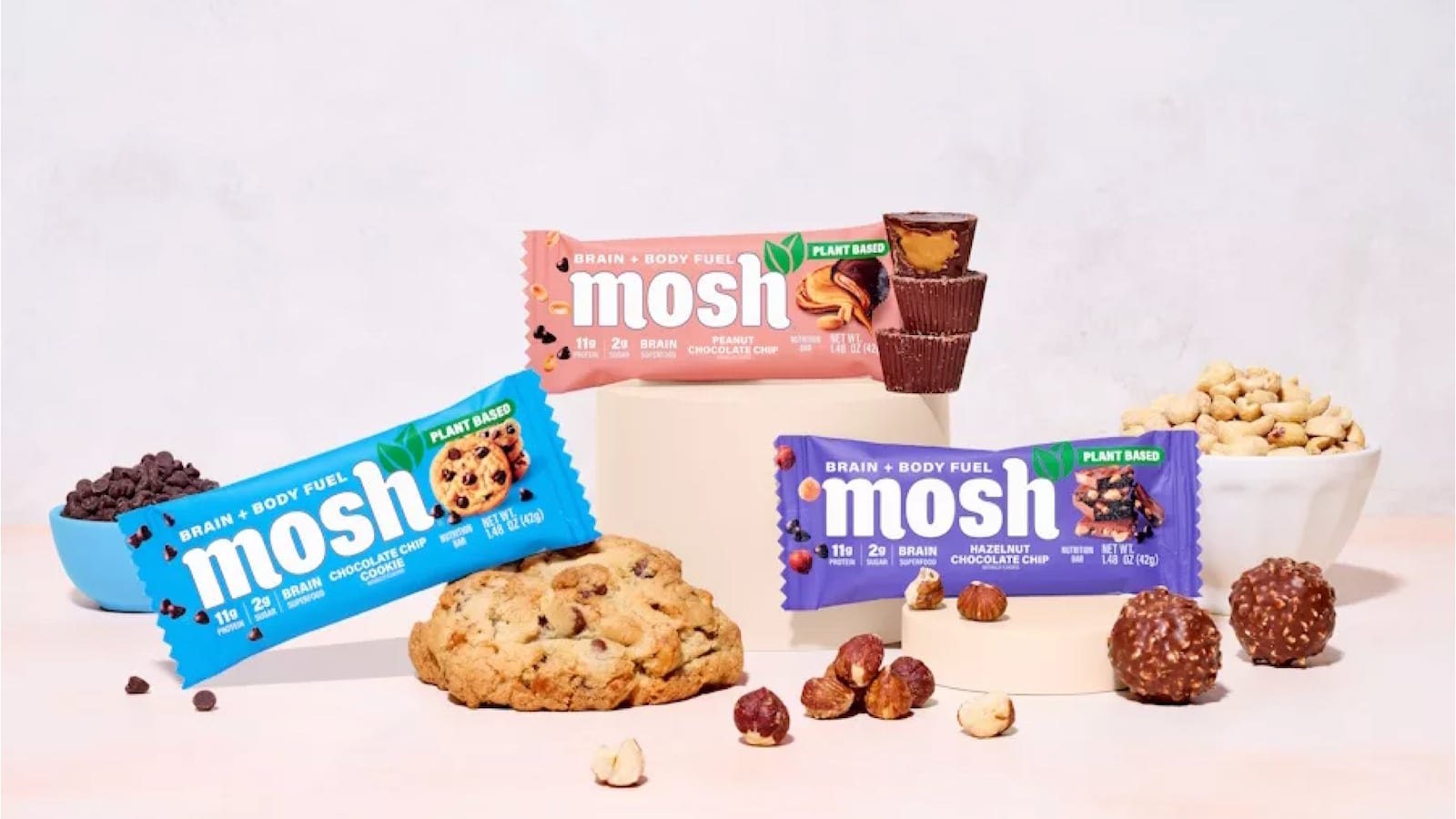 Three MOSH protein bars in Chocolate Chip Cookie, Peanut Chocolate Chip, and Hazelnut Chocolate Chip flavors, surrounded by their real ingredients including cookies, peanut butter cups, hazelnuts, and chocolate chips."