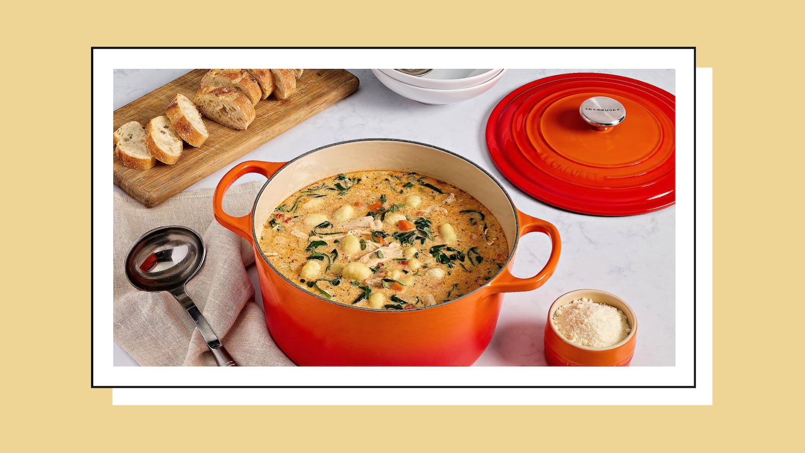Orange Le Creuset Dutch oven filled with creamy gnocchi soup, surrounded by sliced bread, grated cheese, and a ladle, styled on a kitchen table.