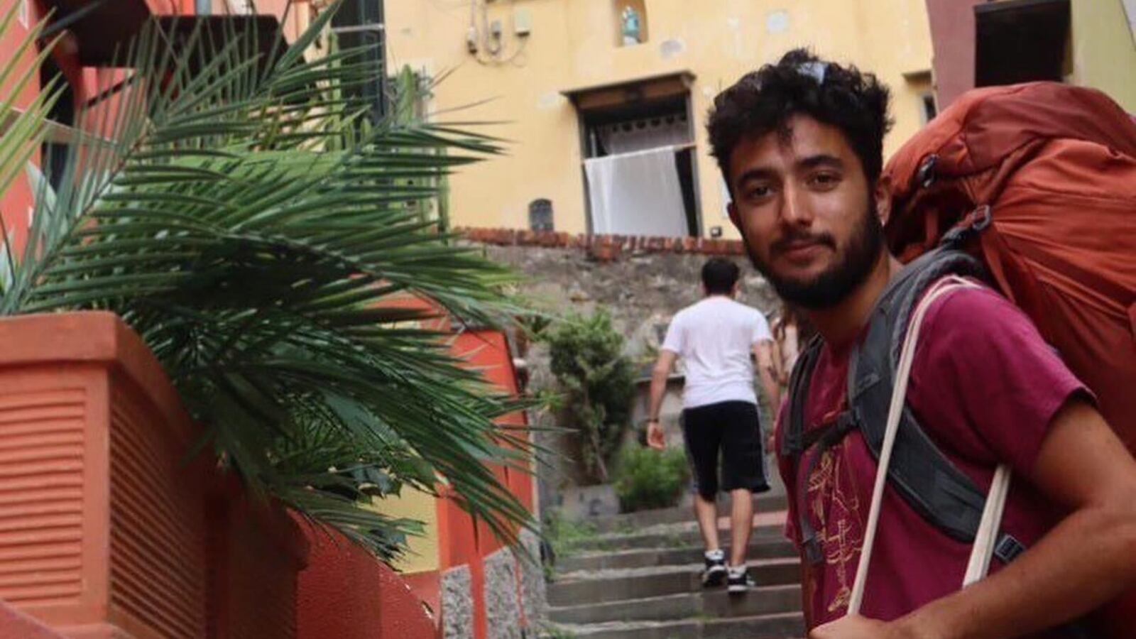 An image of Hersh Golberg-Polin, wearing an orange backpack, in front of a yellow building and stone steps.