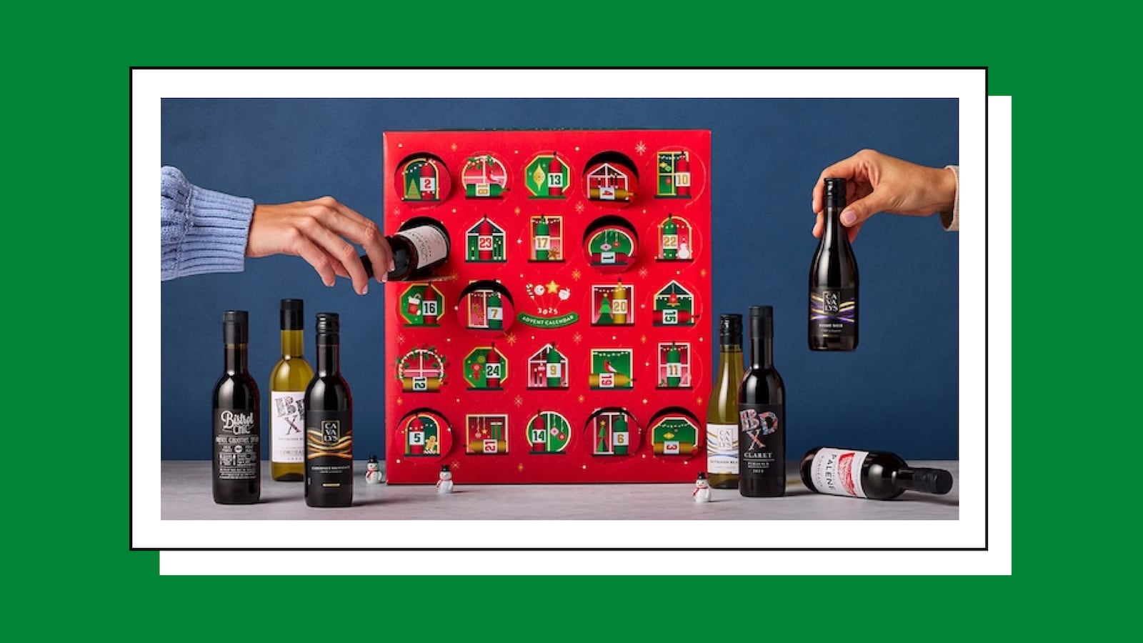 A red wine advent calendar box with 24 numbered doors, some opened to reveal mini wine bottles, while two hands hold different bottles. Several other small wine bottles stand in front of the calendar alongside tiny snowman decorations
