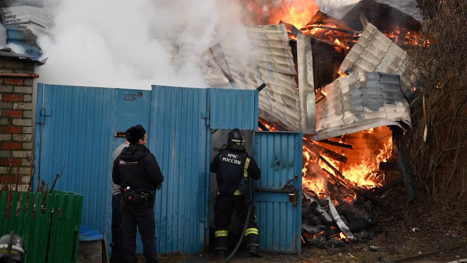 Emergency personnel work to put out a fire before a blue building.