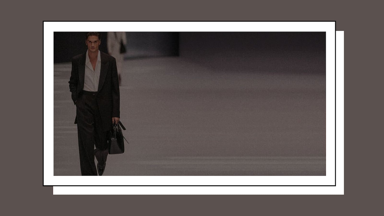 Male model in a dark oversized suit walking on a fashion runway holding a black handbag, exuding confidence and style in a minimalist, high-fashion setting.