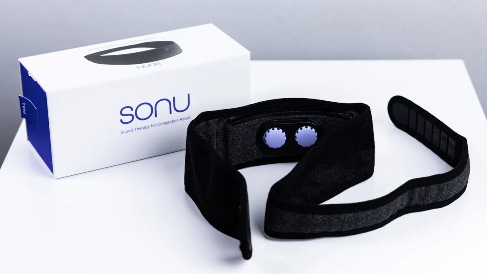 SoundHealth's new SONU Headband. The headband and packaging are on a white table. The headband is black with two purple circles. The packaging is a white and purple with the word SONU on it and a picture of the headband.