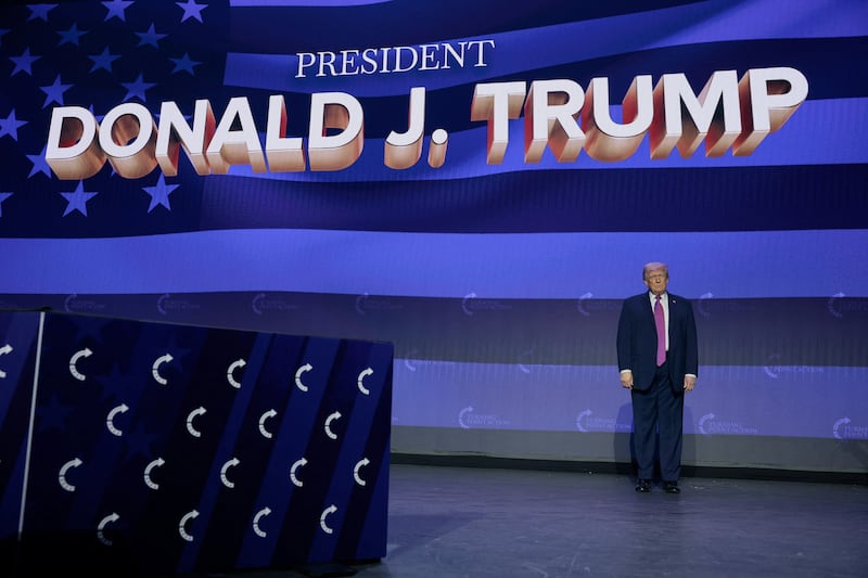 U.S. President Donald Trump stands on stage in front of a screen showing the American flag and his name in large letters.