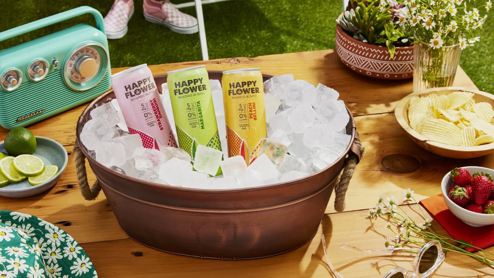 Three Happy Flower cans in an ice bucket on a bench surrounded by a bowl of strawberries, a plate of limes, a radio, a bowl of chips, and a vase of flowers.