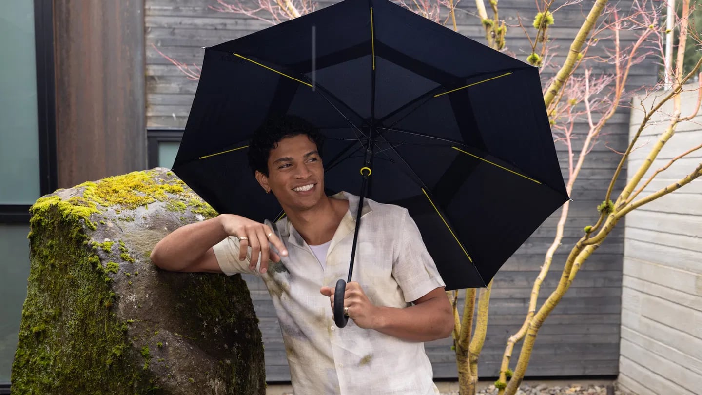 Man holding SHED RAIN umbrella and smiling while leaning on a rock