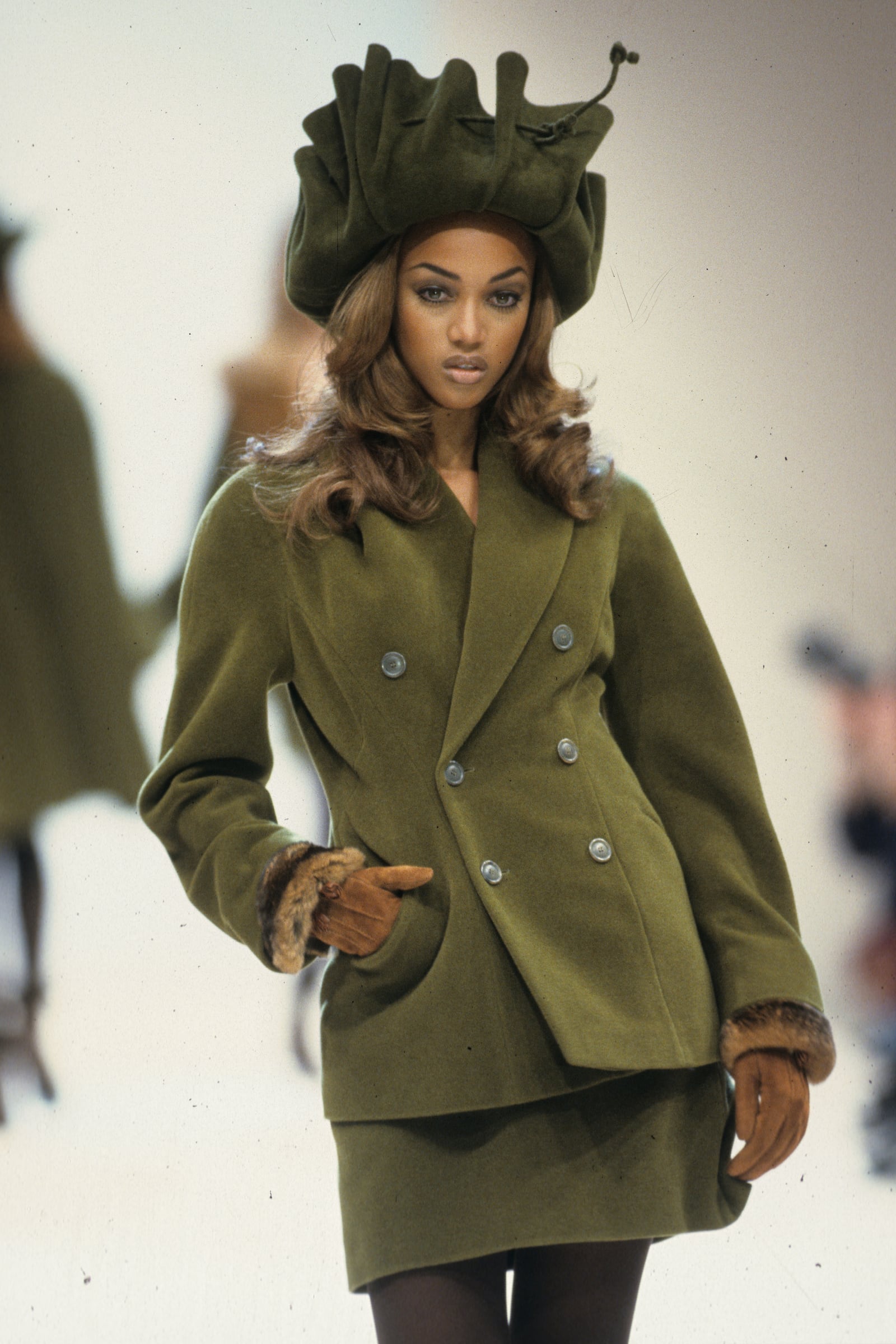 Outtake; Also appeared in WWD 4/29/1992 p.8; Model Tyra Banks wears a green double-breasted jacket over a wool mini-skirt in a matching hue along with boots and a contemporary take on the trapper's hat on the runway for designer Jil Sander in Milan..Article title: Bread-and-Butter