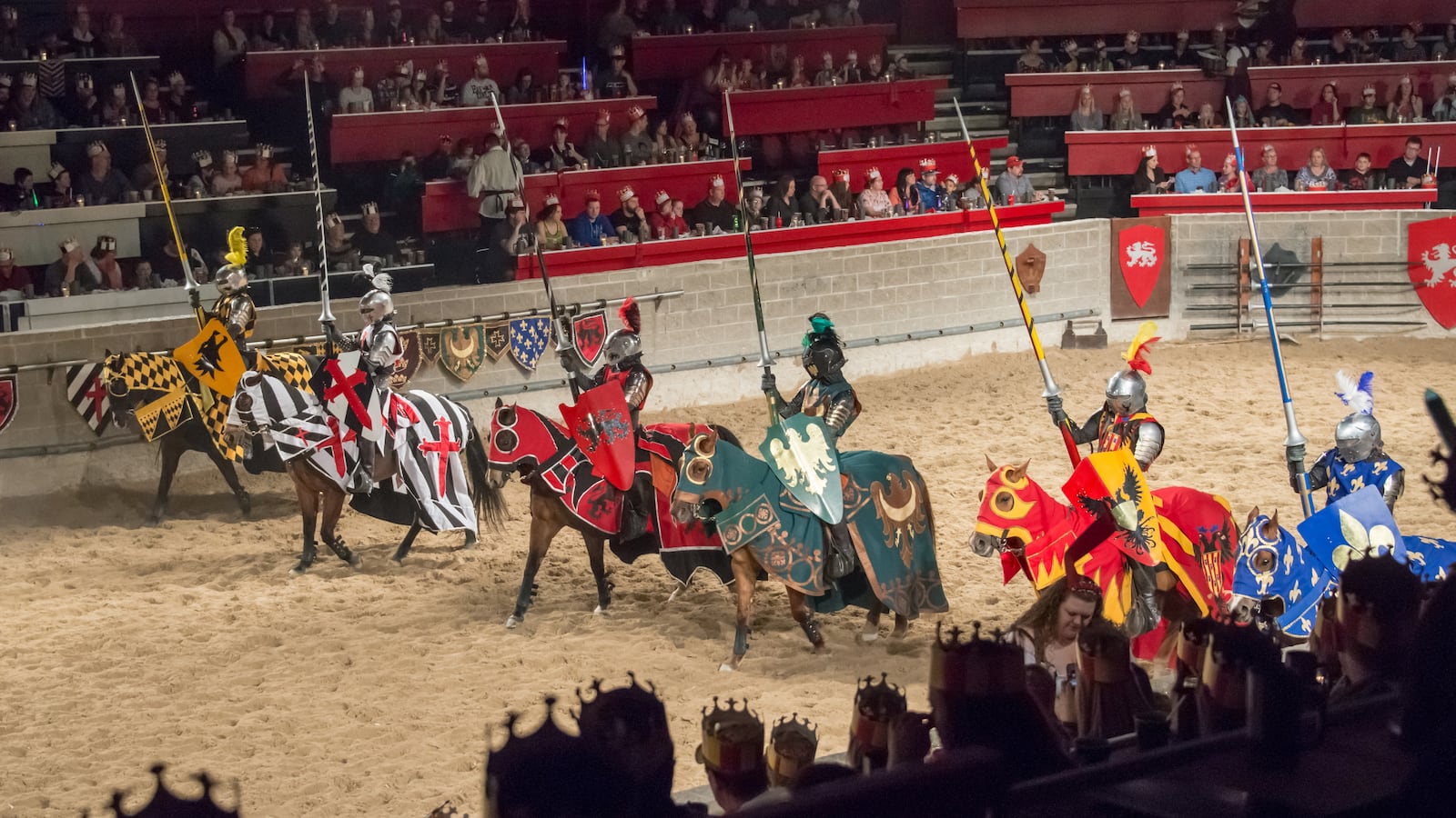 Medieval Times Restaurant: Six horsemen at a jousting event, dressed as medieval knights holding lances, lined up in a stadium full of spectators, waiting to begin the show.