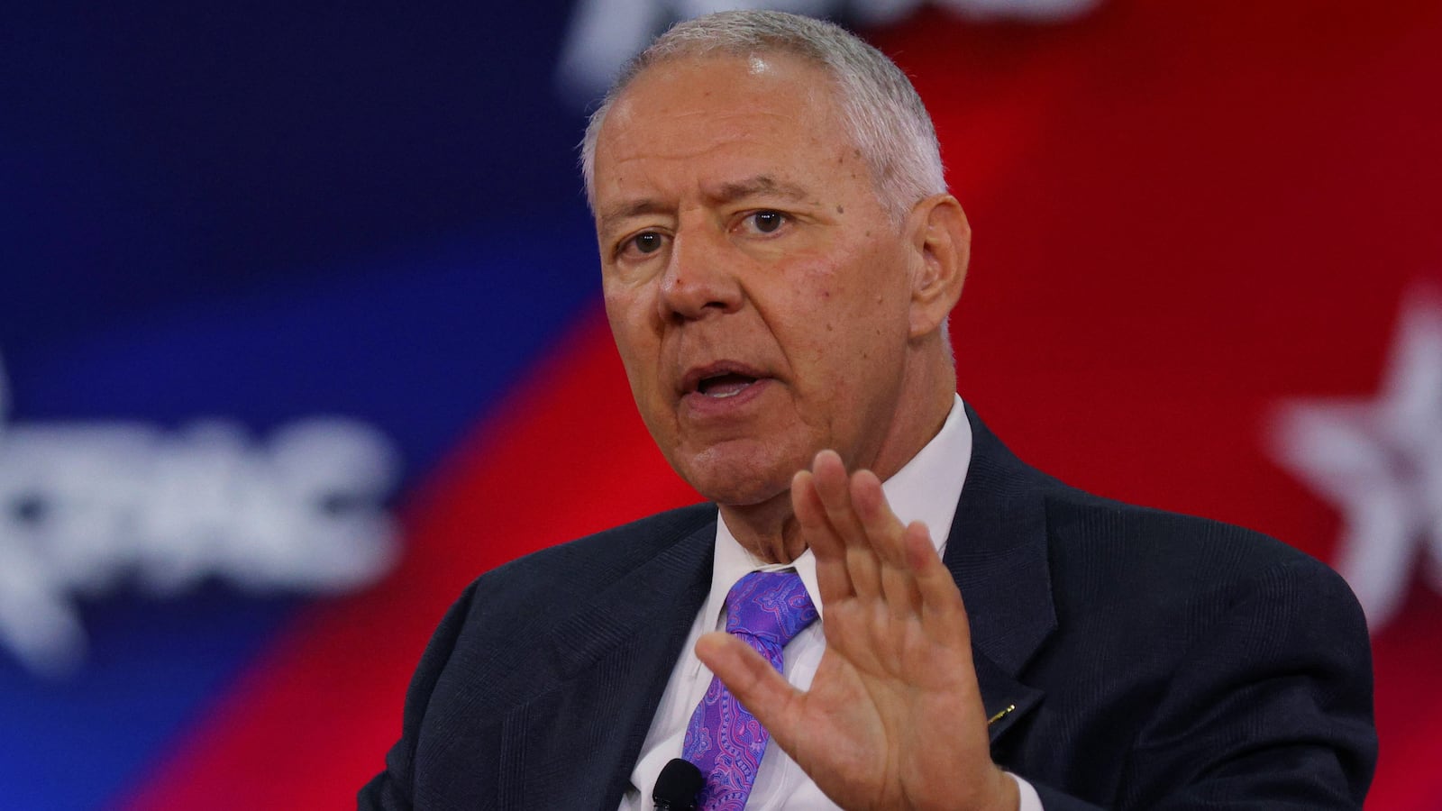 Ken Buck speaks on stage with a hand raised at an event.