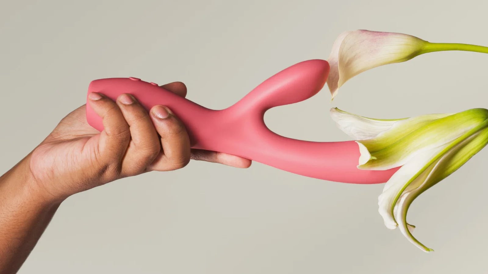 A hand grips a coral-pink rabbit vibrator as it meets a white calla lily, the two heads of the toy emerging from the flower's petals against a soft gray background.