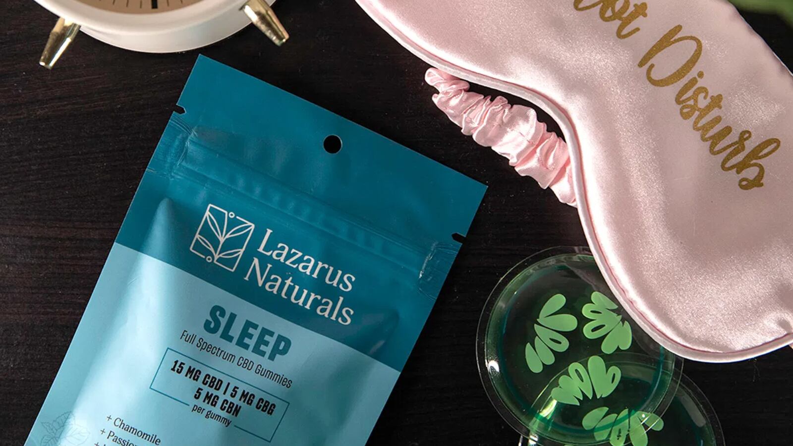Lazarus Naturals's Sleep Gummies on a table next to a glass vase, pink sleep mask, and clock.