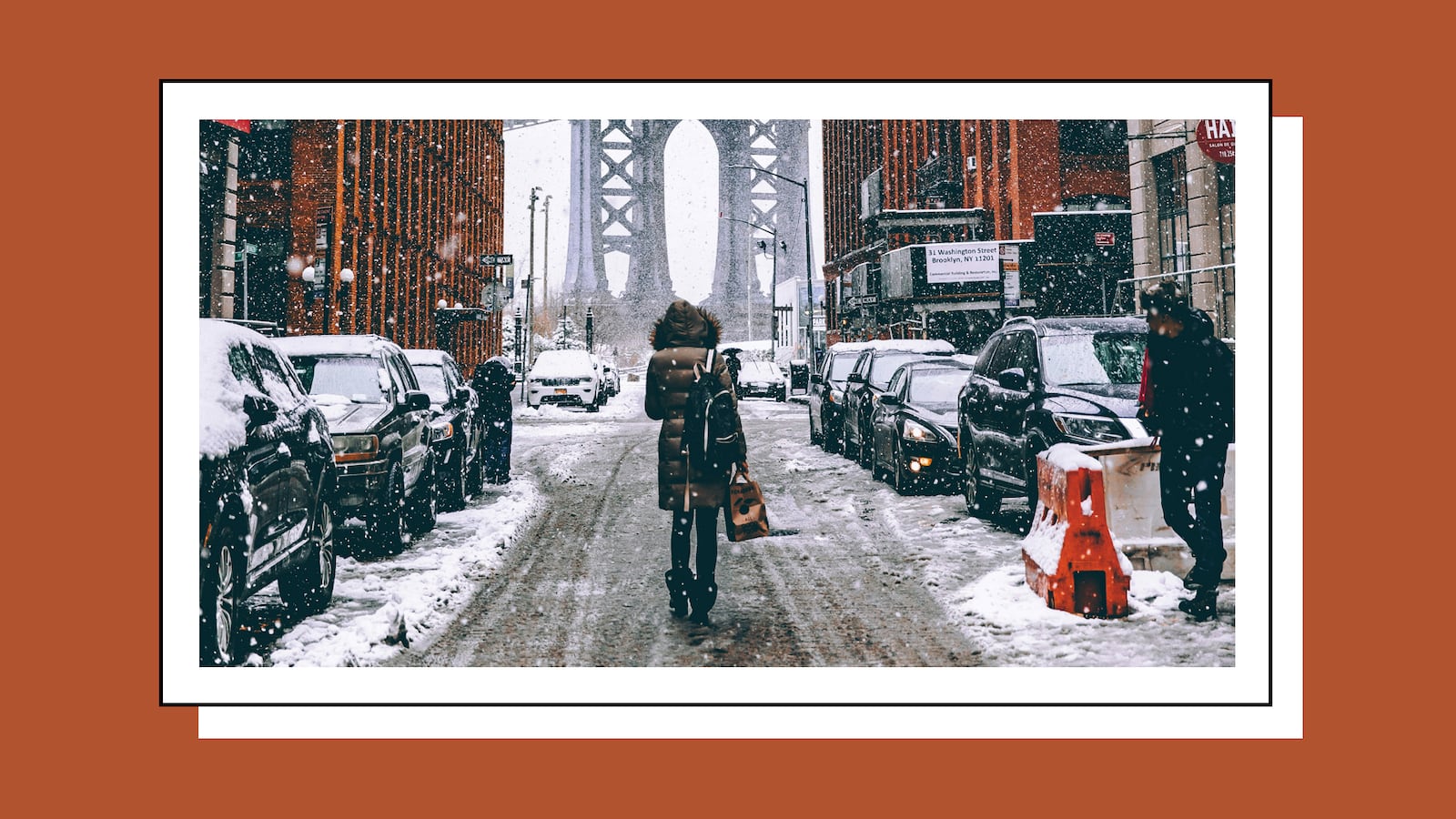 Woman wearing a winter jacket walking down a snowy street with cars on both sides of her.