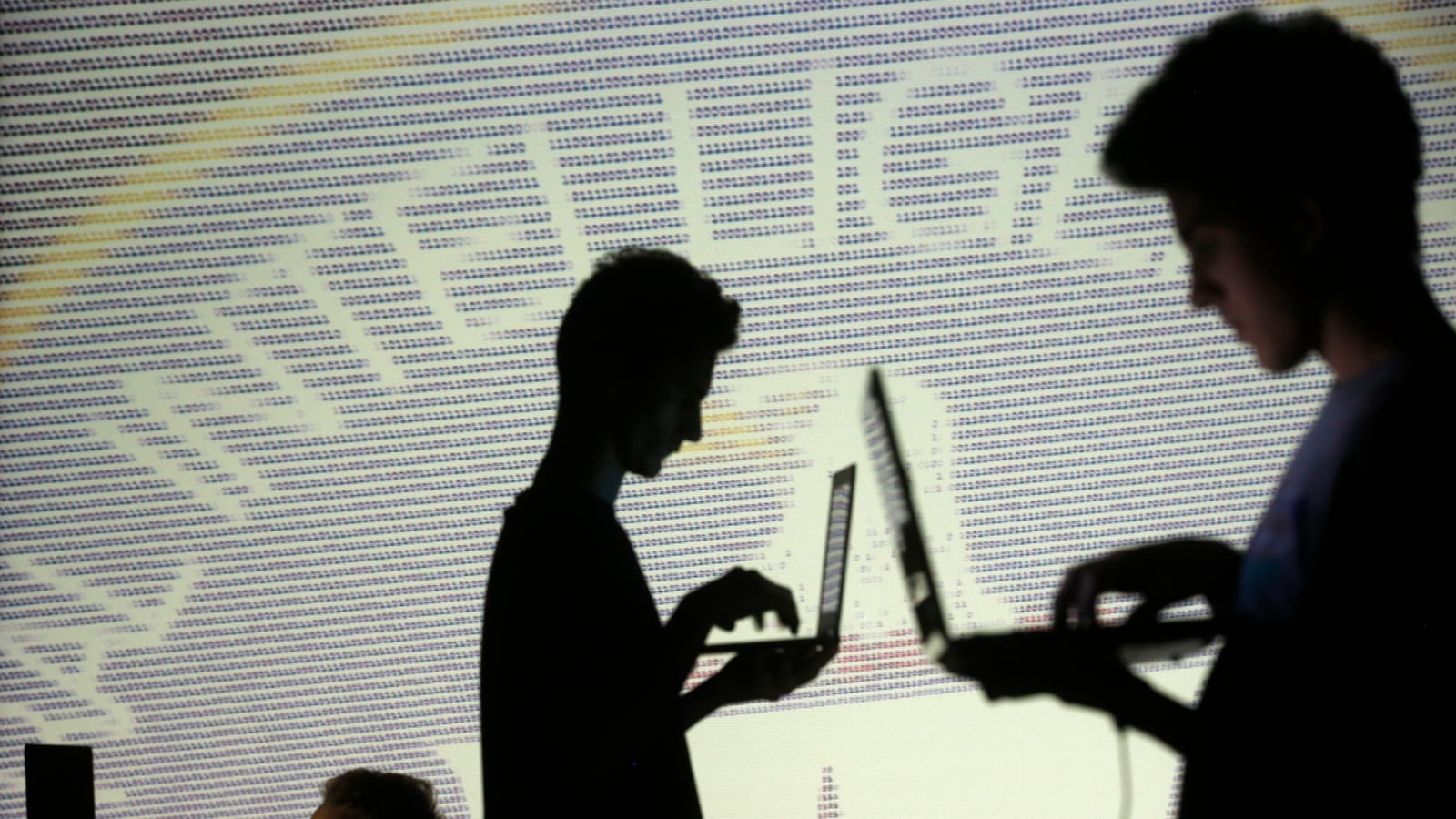 People are silhouetted as they pose with laptops in front of a screen projected with binary code and a Central Inteligence Agency (CIA) emblem.