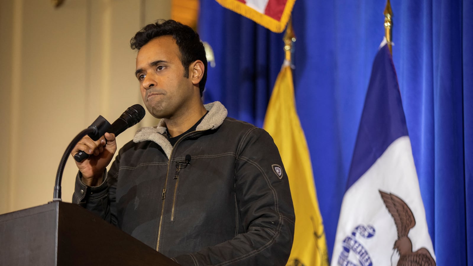 Republican presidential candidate Vivek Ramaswamy frowns while giving remarks at a podium. He's standing in front of US and Iowa state flags.