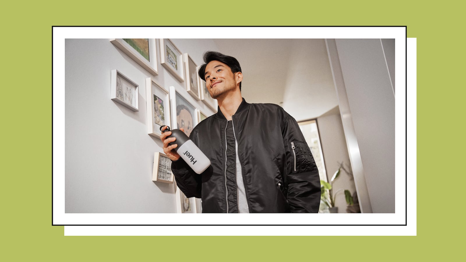 A man smiling confidently while holding a Huel-branded bottle, standing in a stylish, well-lit interior with framed wall art in the background.