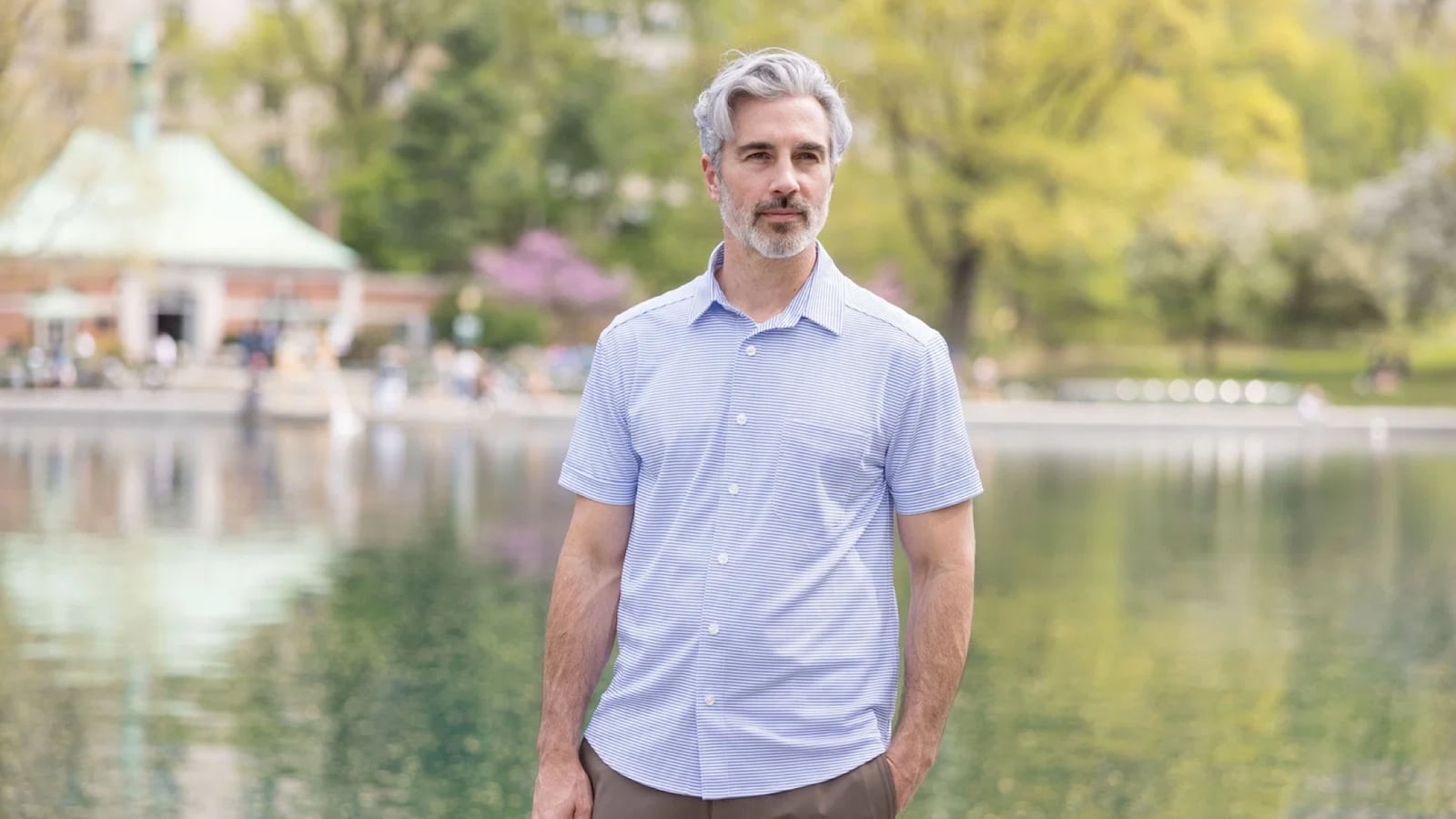 Man wearing a light blue striped short-sleeve button-down shirt, standing in front of a reflective pond in a park setting.