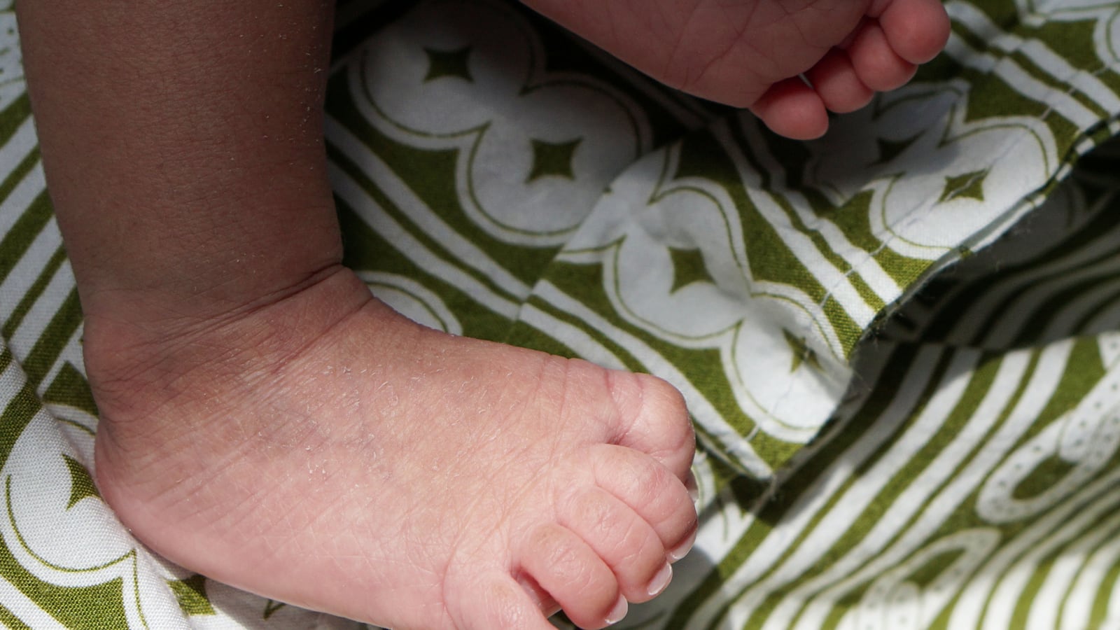 A heartwarming close-up shot focusing on the delicate, wrinkled feet of a sleeping infant, resting comfortably on a richly patterned fabric of olive green and white stripes and clover-like designs und