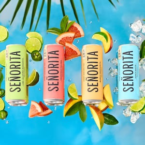 Four canned beverages–light green, peachy pink, tangerine, and sky blue– sit on a mirror that's reflecting clear blue sky and spiky palm leaves. Around the beverages are ice and fruits: lime, mango, grapefruit, jalapeño, mint – the flavors of the THC beverages.