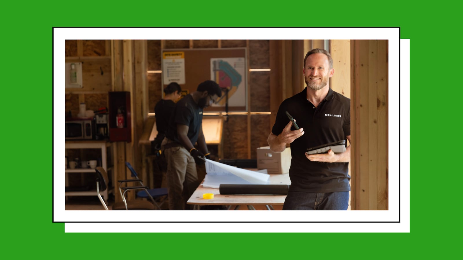 Small business contractor using a tablet and phone at a job site while team members review blueprints, illustrating digital tools for managing business operations.