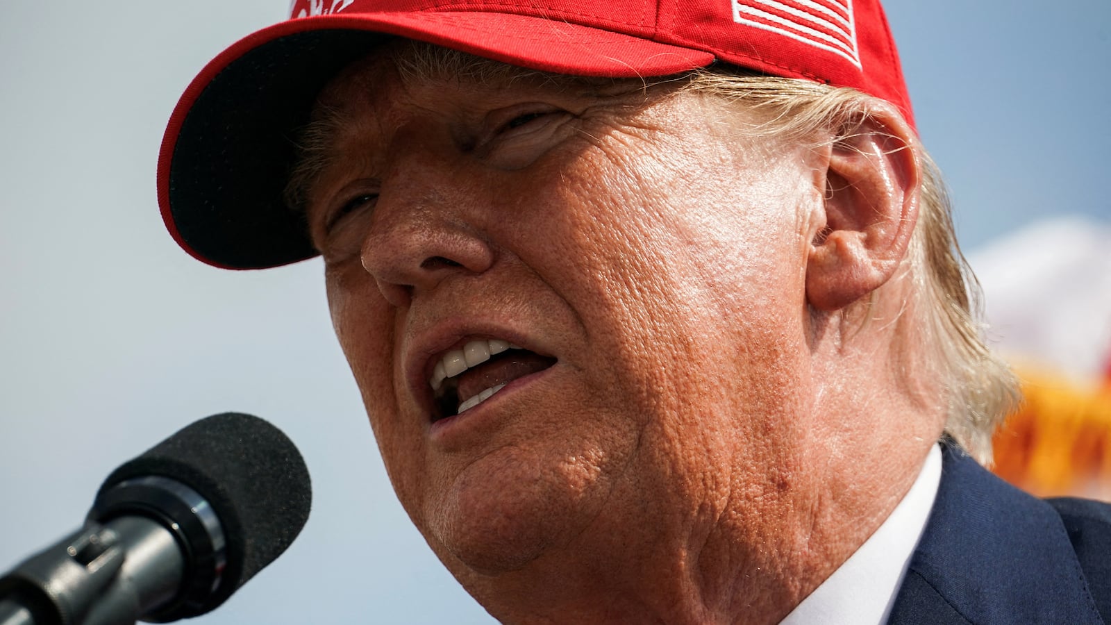 Donald Trump, wearing a red “Make America Great Again” hat, speaks on stage.