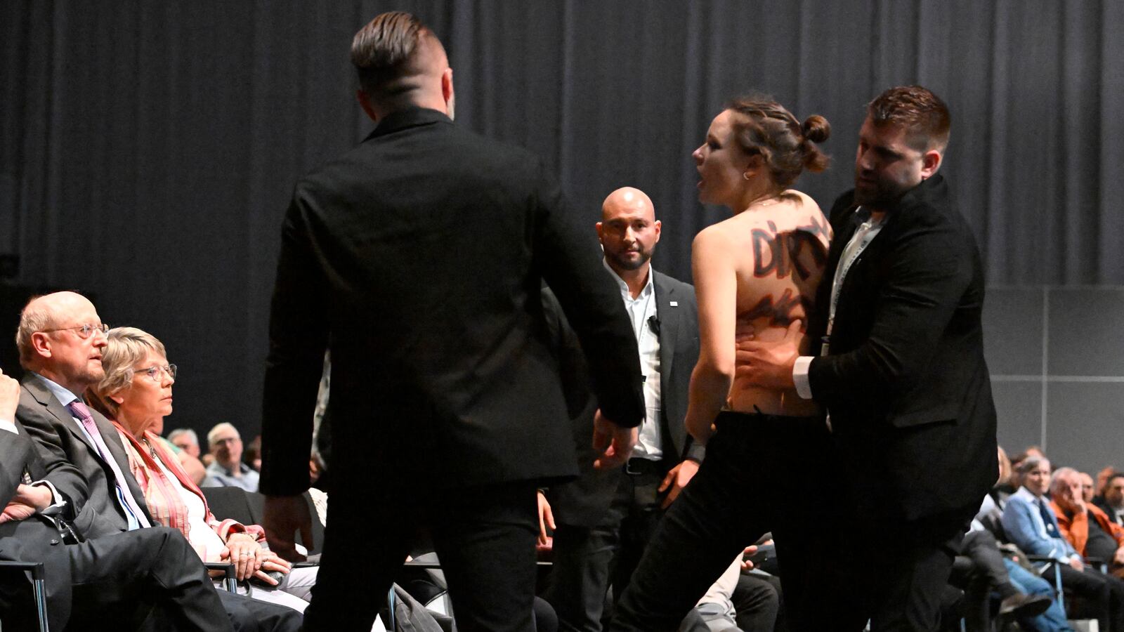 A topless activist is escorted away as she demonstrates against Volkswagen's ongoing production of cars with combustion engines using fossil fuels during the annual general meeting of German carmaker Volkswagen (VW) in Berlin on May 10, 2023.