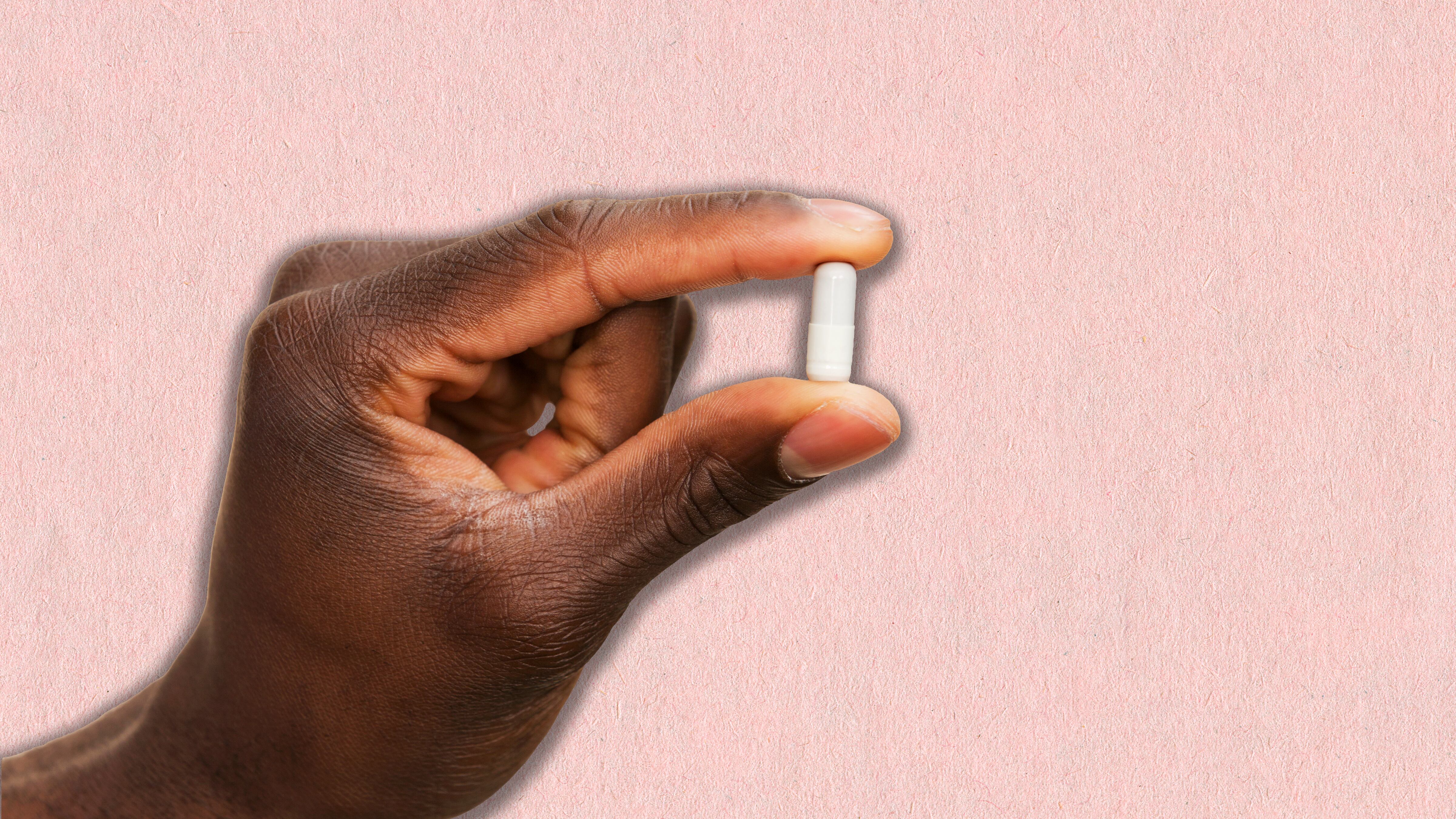 a hand holding up a small white supplement pill over a pink background