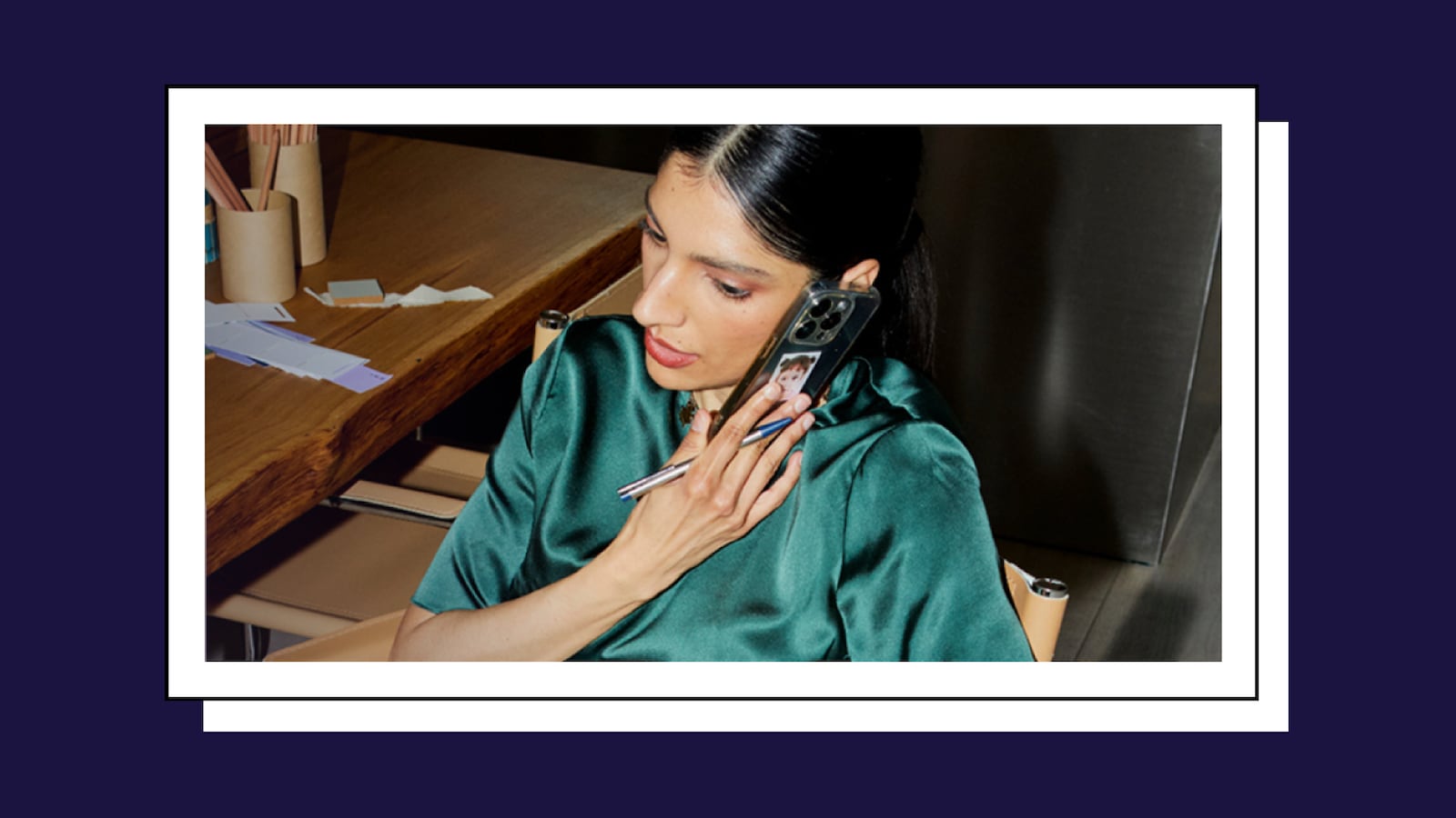 Woman in a green satin blouse holding a phone to her ear at a wooden desk, with pens, papers, and a mug of pencils nearby.