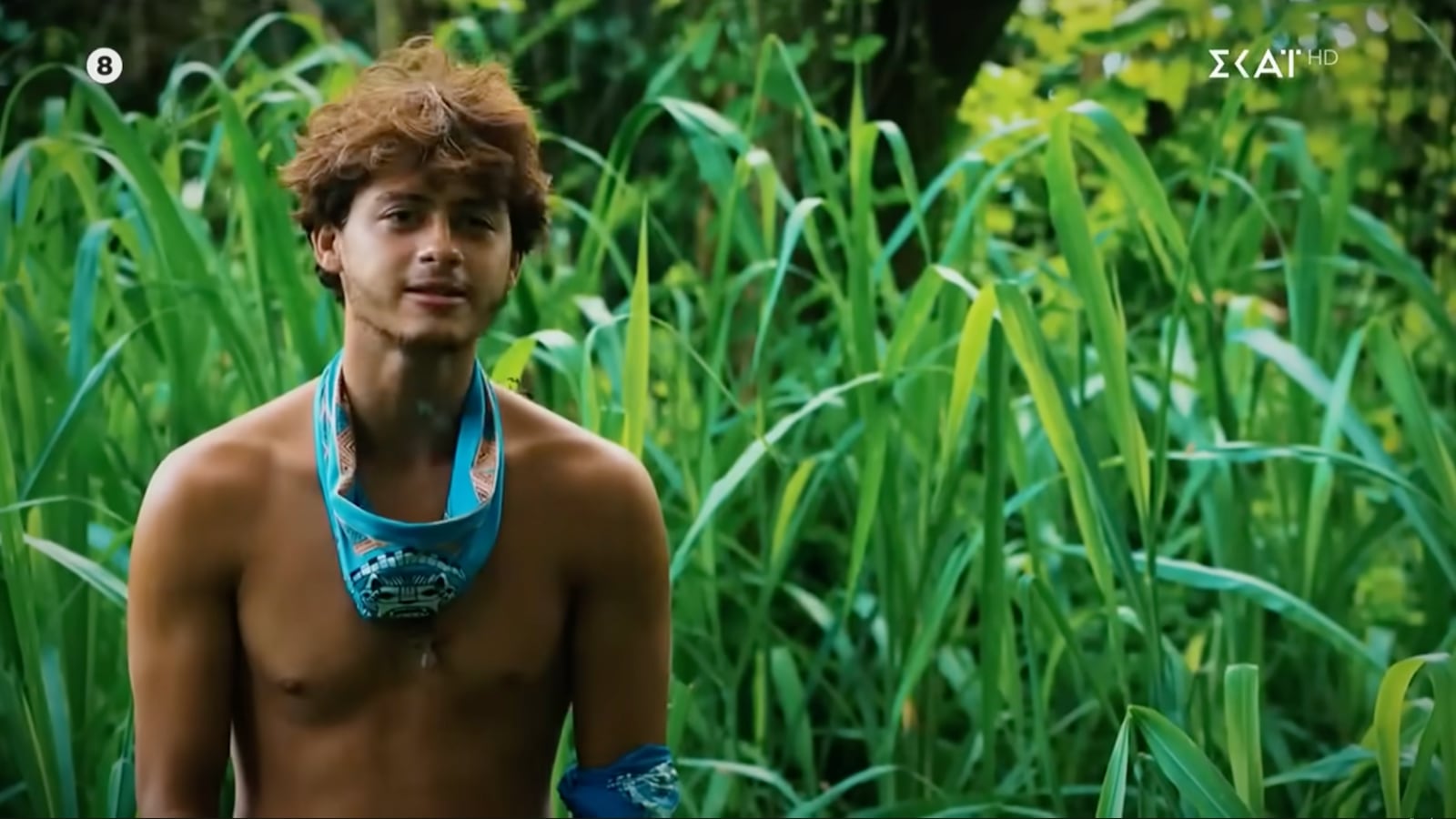 A man stands among tall grasses wearing a bandana around his neck.