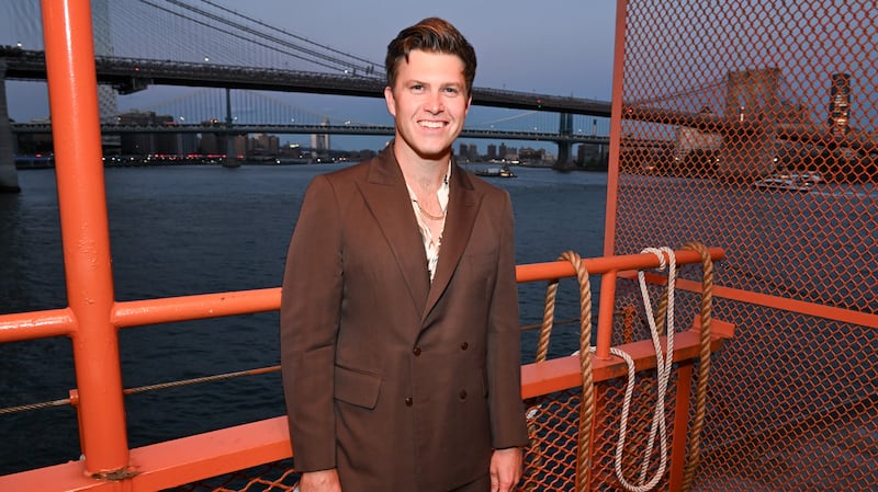 Comedian Colin Jost wearing a brown suit and standing and smiling aboard the Staten Island Ferry that he owns.