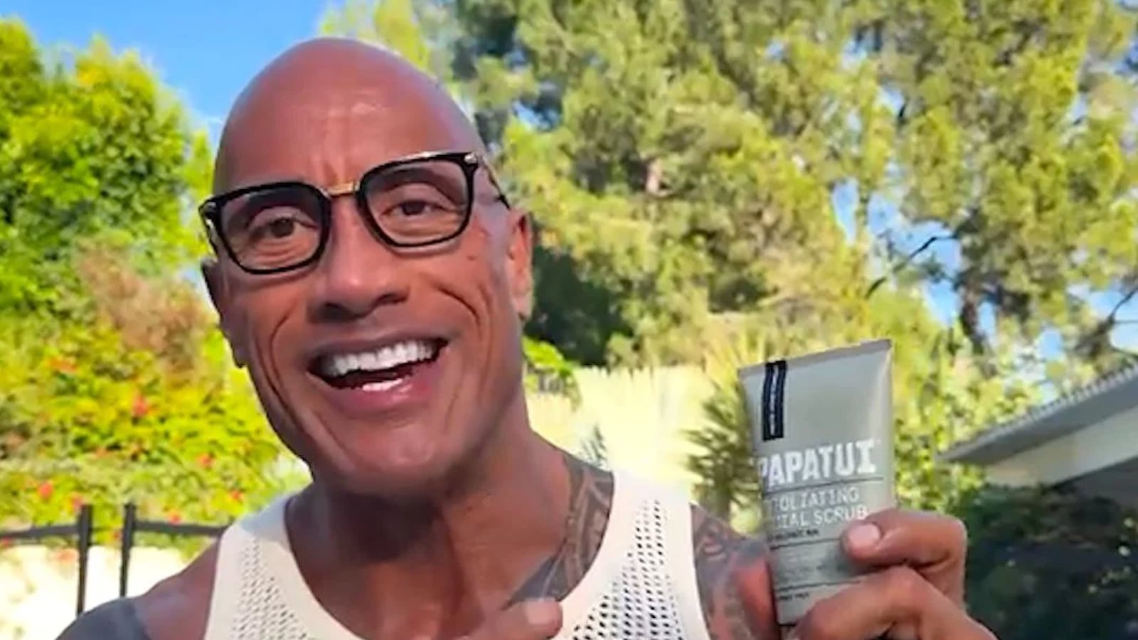 Dwayne "The Rock" Johnson holding PAPATUI Exfoliating Facial Scrub tube outdoors, smiling.